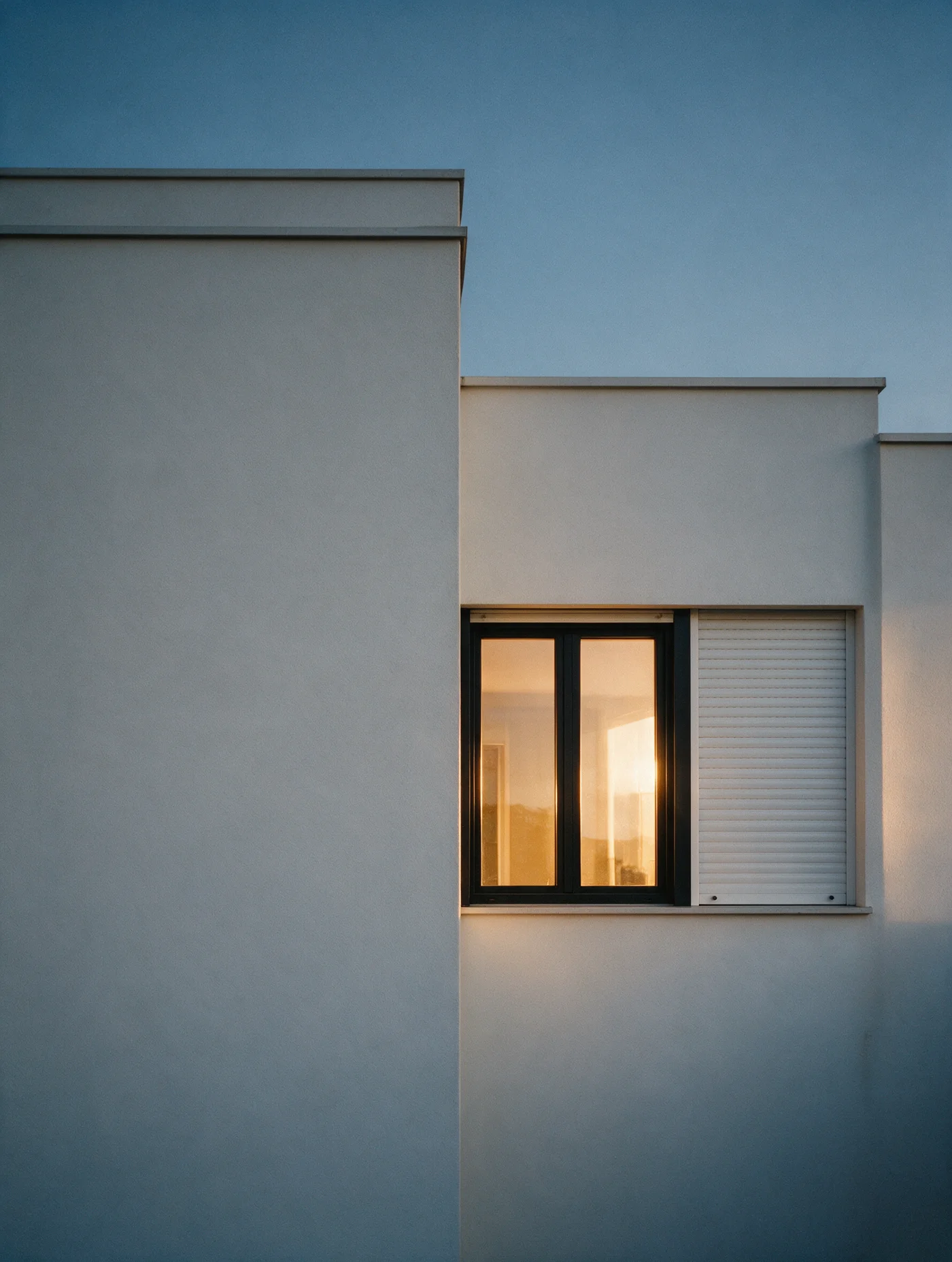 A modern villa facade at dusk with an aluminum window system and a roller shutter, warm interior light glowing through the glass